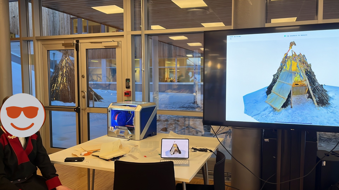 Photograph of a Sámi duodji, or handicraft, student sitting at a table with a 3D printer, a laptop, and a tv showing a 3D scanned model of a lavvo, or traditional Sámi dwelling. Outside through the window sits the actualy Sámi dwelling that was scanned and you can see the scan is very lifelike.
